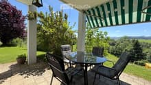 Balcony with a glass table and four chairs, overlooking a green landscape with trees and hills.