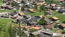 Aerial view of a small village with numerous houses, green lawns, and scattered trees. The houses have varied roof styles and colors, and the area is surrounded by mountains.