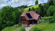 A two-story wooden house with a red-tiled roof, surrounded by lush greenery and hills.