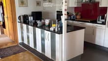 Modern kitchen with white cabinets, black countertops, wooden herringbone flooring, and various appliances including a coffee maker, microwave, and oven. A pet bowl is visible on the floor.