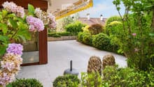 Well-maintained garden with lush greenery, flowering shrubs, decorative statues, and a covered patio area with a glass door leading inside.