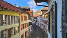A narrow cobblestone street lined with colorful, multi-story buildings with traditional European architecture, featuring wooden shutters and tiled roofs.