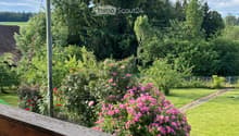 Wooden balcony overlooking a lush garden with pink flowers, green grass, and tall trees under a partly cloudy sky.