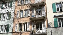 Multi-story residential building with balconies, green shutters, and a tree in front.