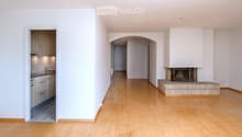 Empty living space with light wooden flooring, white walls, and a fireplace with a tiled surround. A small kitchen area is visible through an open doorway.