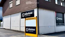 Two-story building with brown wooden facade, multiple windows, and signage for "bikebox.ch" and "SCHMERIKON" on the walls. A yellow signpost in front displays the address "Hantteilstrasse 2".