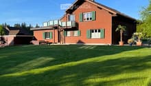 Two-story red brick house with green shutters, large windows, and a balcony on the upper level. The house is surrounded by a well-maintained lawn and features potted plants near the entrance.