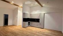 Empty room with wooden flooring, white walls, and a small kitchen area with cabinets and appliances.