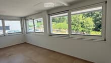 Empty room with tiled floor, white walls, and large windows offering a view of greenery outside.
