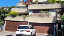 Modern two-story house with a flat roof, large wooden garage doors, and a white SUV parked in front. The house features a raised ground floor and is surrounded by greenery and plants.