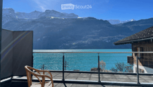 Balcony with a view of a lake and mountains, featuring a single wicker chair and a metal railing.