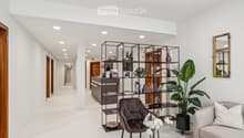 Modern, minimalist waiting area with white walls, a grey sofa, a dark brown leather chair, a large potted plant, a metal shelving unit with books and decorative items, and a reception desk in the background.