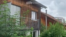 Two-story wooden building with a flat roof and balcony, surrounded by greenery.