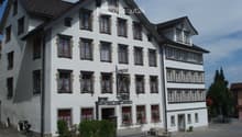 A three-story white building with red-framed windows and decorative trim, featuring a sign reading "Gasthof zur Krone" and a small outdoor seating area with tables and benches.