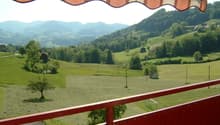 Balcony with red wooden railing, overlooking a scenic valley with green fields, scattered trees, and distant mountains.