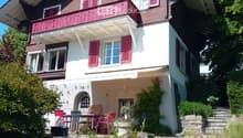 Two-story house with red brick and white siding, multiple windows with shutters, a balcony on the upper level, and a small patio area with outdoor furniture.