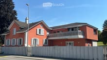 Two-story red house with white shutters, balcony on the upper floor, surrounded by a concrete fence.
