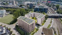 Aerial view of a modern residential complex with solar panels on the rooftops, surrounded by greenery, parking areas, and nearby urban infrastructure including roads, train tracks, and other buildings.