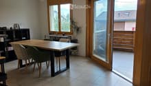 Bright dining area with tiled floor, large wooden dining table, four chairs, a small shelf with decorative items, and a potted plant near the window. A sliding glass door leads to a balcony.