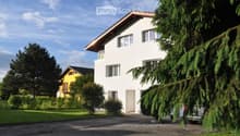 2 story house, white exterior, brown roof, balcony, driveway, car parked, trees, bushes, yellow house in background