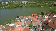 Aerial view of a town with numerous houses, red-tiled roofs, and a lake in the background.