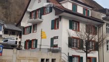 A multi-story residential building with white walls, green shutters, and red trim around windows. Features include a raised ground floor, a stone staircase, and a small balcony on the upper level.