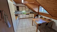 Bright kitchen with white cabinets, wooden ceiling beams, tiled floor, and a dining area with a wooden table and chairs. The kitchen features a refrigerator, dishwasher, and various small appliances on the counter.