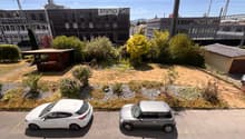 A small garden area with a wooden gazebo, surrounded by bushes and trees, with two parked cars in the foreground.
