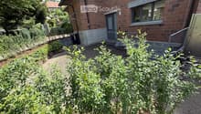 A small garden area in front of a brick building, featuring a gravel pathway, green shrubs, and a small bench. The building has a modern design with large windows and a raised entrance.