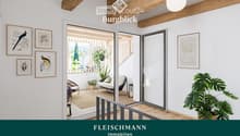 Bright living room with white walls, wooden ceiling beams, large windows, and a view of the balcony. Features a sofa, small table, chairs, and decorative plants.