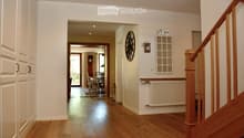 Spacious hallway with wooden flooring, white walls, and a staircase leading upwards. Features a radiator, a wall-mounted clock, and a framed picture. A dining area is visible in the background with a table and chairs.