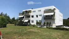 Modern white multi-story building with balconies and greenery, surrounded by a grassy lawn and trees.