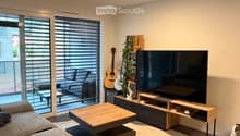 Modern living room with a grey sofa, wooden coffee table, large flat-screen TV, and a guitar leaning against the wall. The room features large windows with blinds and a view of a balcony area.