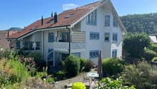 Two-story house with white exterior, red-tiled roof, multiple windows, and a small balcony. The surrounding garden features a patio with outdoor furniture and plants.