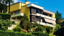 A modern two-story house with yellow and white exterior, featuring balconies, large windows, and surrounded by lush greenery and trees.