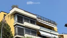 Multi-story residential building with balconies, large windows, and surrounding greenery including palm trees and shrubs.