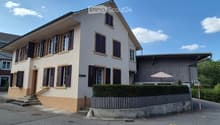 Two-story house with white walls, dark shutters, and a raised ground floor accessed by steps.