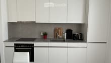 Modern kitchen with white cabinets, grey countertops, and stainless steel appliances. Includes a sink, oven, coffee maker, teapot, and a small potted plant on the counter.