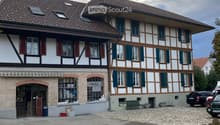 Two-story traditional building with white and brown timber framing, multiple windows with shutters, and a tiled roof. A small entrance area with a bench and potted plants is visible.