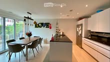 Modern kitchen with white cabinets, a central island, a dining table with chairs, and a vase of flowers. The space features recessed lighting and a pendant light above the island.