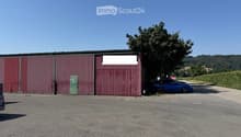 A large industrial-style building with red corrugated metal walls and a green door, situated in a rural area with a tree and a blue car parked nearby.