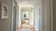 Elegant hallway with herringbone wooden flooring, white walls, and multiple doors leading to different rooms. A framed artwork is displayed on the left wall, and a small rug lies in the center of the hallway.