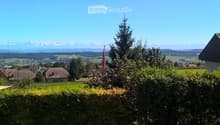 A scenic outdoor view with a hedge in the foreground, houses and trees in the middle ground, and a panoramic mountain range in the background under a clear blue sky.