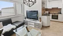 Modern, open-plan living and dining area with a black leather sofa, glass coffee table, flat-screen TV on a white stand, and a compact white kitchen with cabinets, sink, and oven. Large windows offer a city view.