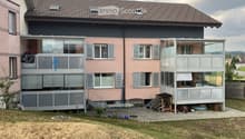 Two-story house with pink and grey exterior, multiple windows, balconies, and a covered patio area. The yard includes a small table, chairs, and bicycles.