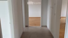 Empty hallway with tiled floor and wooden flooring visible in adjacent rooms, white walls, and a ceiling light fixture.