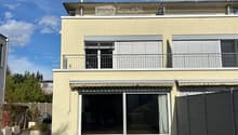 Two-story house with light yellow facade, large windows, balconies, and a covered entrance area.