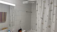 A small bathroom with white tiled walls and floor, featuring a bathtub with a shell-patterned shower curtain, a toilet, a mirror with a light fixture above it, and a towel hanging on the wall. Personal care items are placed on the sink counter.