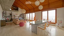 A spacious living room with a high, wooden-beamed ceiling, large windows offering a view, a beige sofa, a colorful ball, a glass dining table with chairs, shelves with books and decorative items, and pendant lamps.