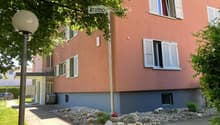 Two-story building with pink facade, white shutters, and a small garden area with rocks and plants.